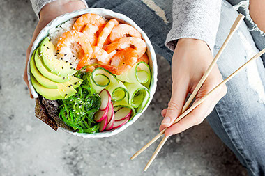 Person holding a shrimp bowl with seaweed, avocado, cucumber, radish, sesame seeds.