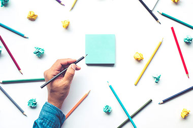 Hand writing on a notepad with colored pencils all around.