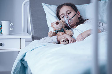 Girl in bed with breathing mask on while holding a teddy bear.