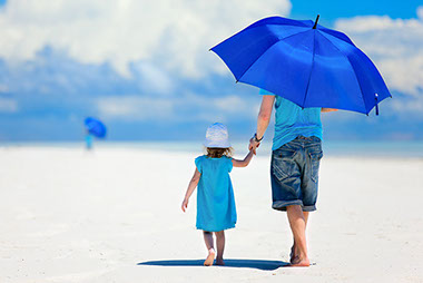 Melanoma and skin cancer awareness. Image of father and child walking on the beach with a big blue umbrella.