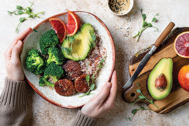 Hands holding tempeh buddha bowl. Marinated tempeh in marinade with rice, steamed broccoli, avocado.