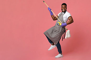 Man wearing apron and cleaning gloves, using a broom stick as a guitar.