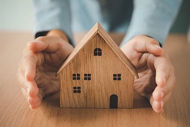 Small wooden home on table with hands protecting it.