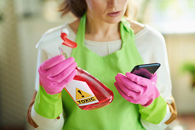 Female wearing cleaning gloves, holding a cleaning bottle with the word "Toxic" on it and looking at her phone in the other hand.