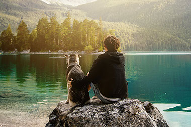 Man sitting on roack along a lake with his dog.