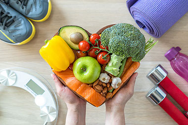 Hands holding a heart shaped plate with fruits and veggies, and exercise gear in the corners.