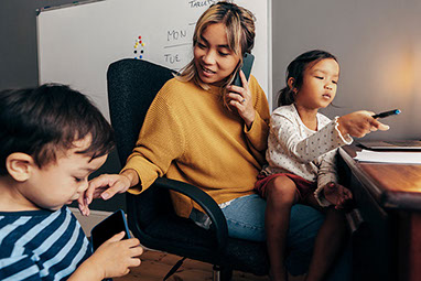 Mother sitting in an office chair, talking on the phone, with 2 young kids