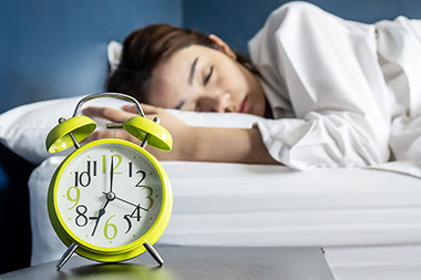 Alarm clock on night stand with female sleeping on bed.