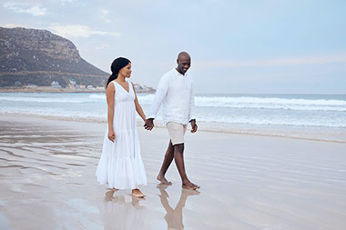 Couple holding hands walking barefoot along the beach with mountains in the background.