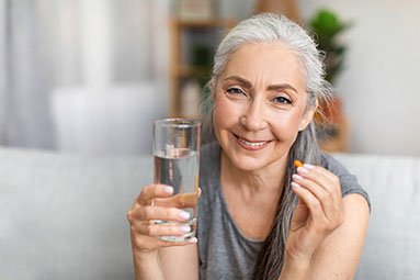 Smiling women holding a glass of water in one hand and a pill in the other hand.