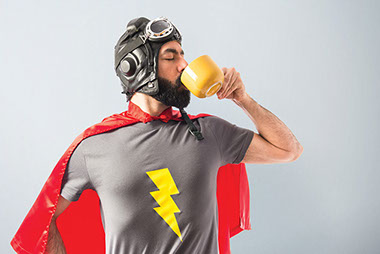 Image of man wearing superhero cape and t-shirt while drinking out of a cup.