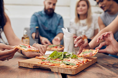 Close up shot of pizza on table, with group of people sitting around and picking up a portion.