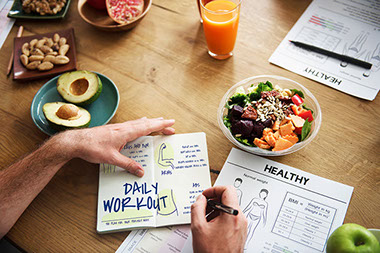 Person writing in fitness notebook with healthy snacks on table.