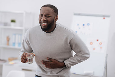 Man holding a glass of water with one hand and holding stomach in pain with the other hand.