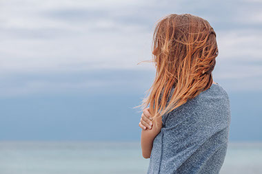 Back of women, wind blowing her hair as she hugs herself.