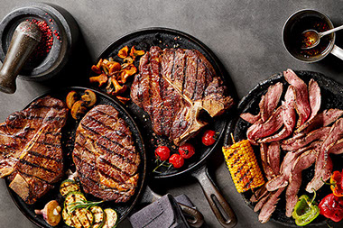 Cooked meats and vegetables in frying pans with seasoning and garnishes.