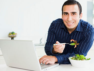 Image of a man with one hand on his laptop computer and the other holding a fork about to take a bite of his healthy salad.